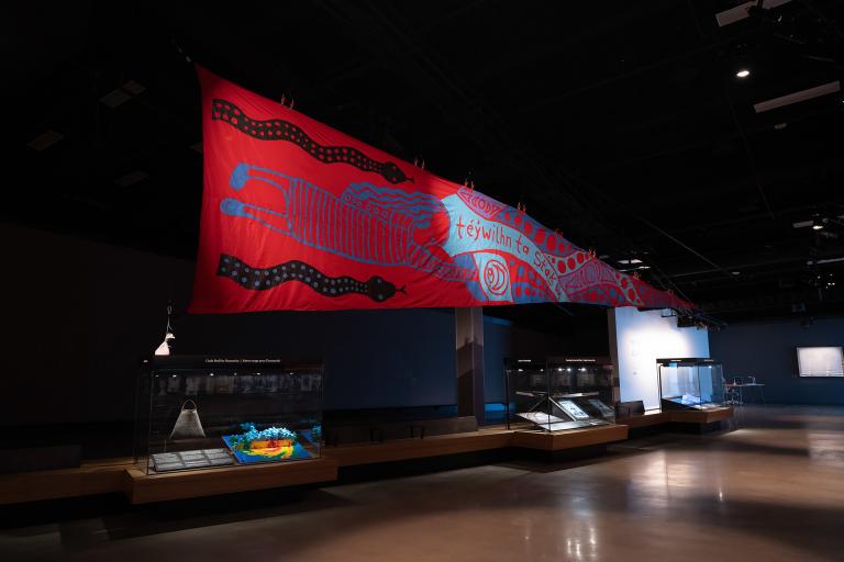 Une grande bannière rouge est suspendue dans la galerie au-dessus de trois vitrines. En français, les mots en squamish sur la bannière signifient : « Protéger cette terre, c’est dans notre sang ». Visibilité masquée.