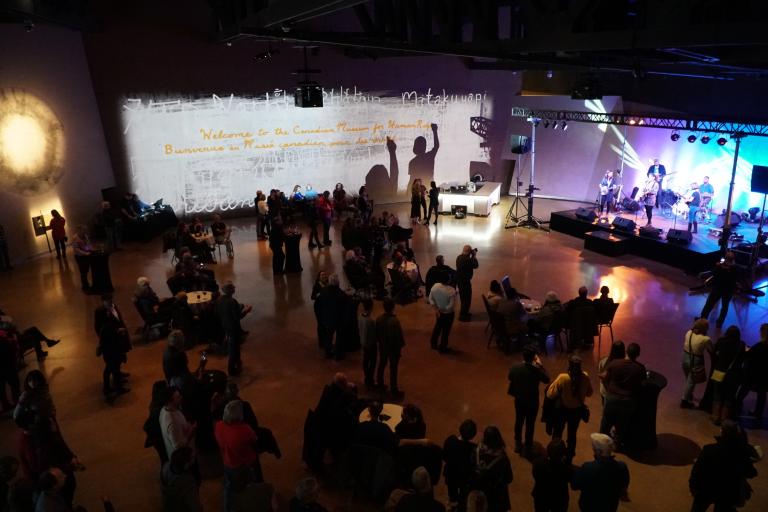 A crowded hall of people watching musicians perform on a brightly lit stage. On the wall in the background, a projected image reads “Welcome to the Canadian Museum for Human Rights.” Visibilité masquée.