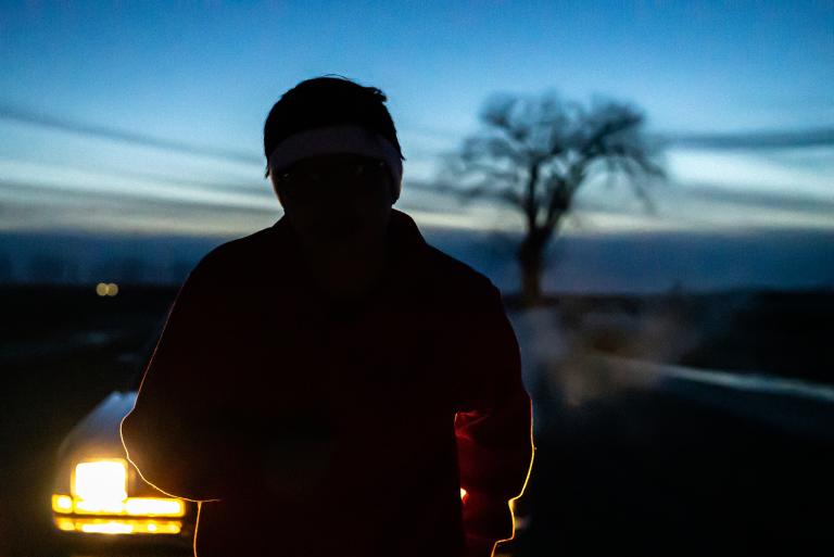 Au premier plan, la silhouette d’une personne qui semble courir vers nous, devant une voiture dont les phares sont allumés. À l’arrière-plan, un ciel bleu pâle avec la silhouette d’un arbre et des nuages longs et fins qui traversent l’horizon. Visibilité masquée.