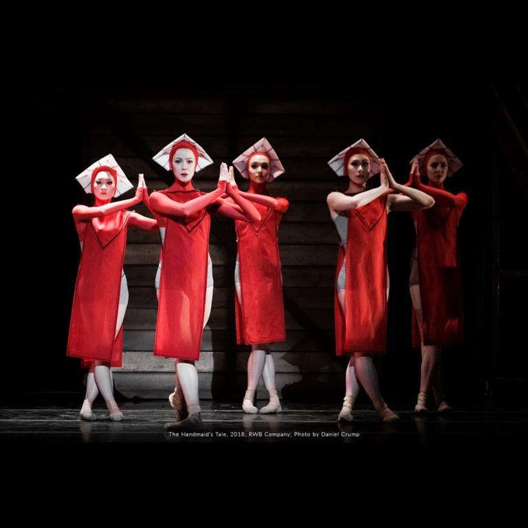 Sur une scène sombre se trouvent cinq danseuses de ballet vêtues de costumes rouges fluides aux genoux et coiffées de chapeaux carrés blancs. Elles regardent vers l'avant, les mains en prière. Visibilité masquée.