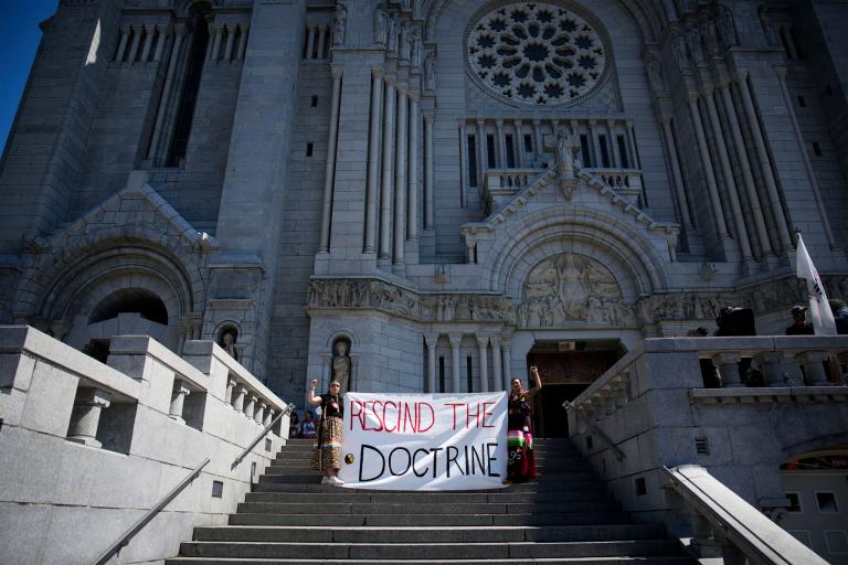 Sur les marches d’une énorme cathédrale, deux personnes portant des tresses et des jupes en ruban lèvent le poing et tiennent une grande bannière en tissu sur laquelle on peut lire en anglais « ABOLIR LA DOCTRINE ». Visibilité masquée.