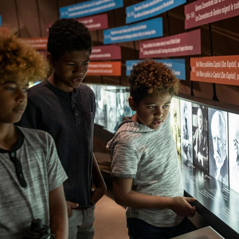 Trois jeunes enfants regardent des photos et un panneau mural. Visibilité masquée.