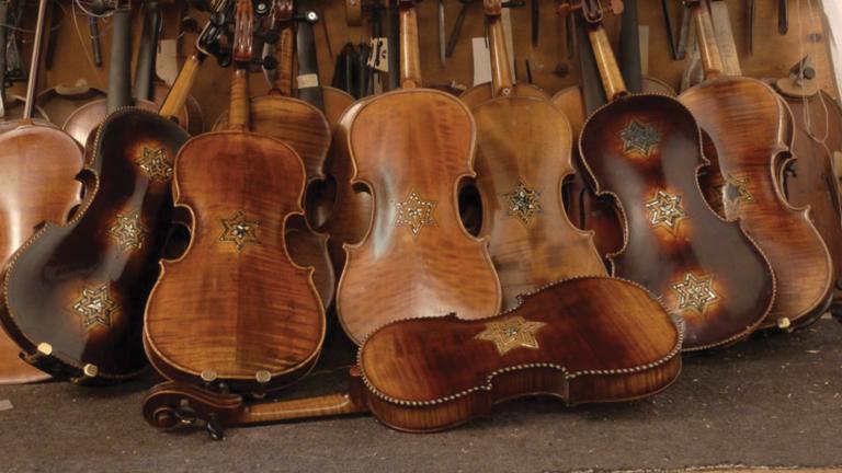 Plusieurs violons restaurés sont présentés appuyés les uns sur les autres dans une pile. Chacun des violons porte au moins une étoile de David. Visibilité masquée.