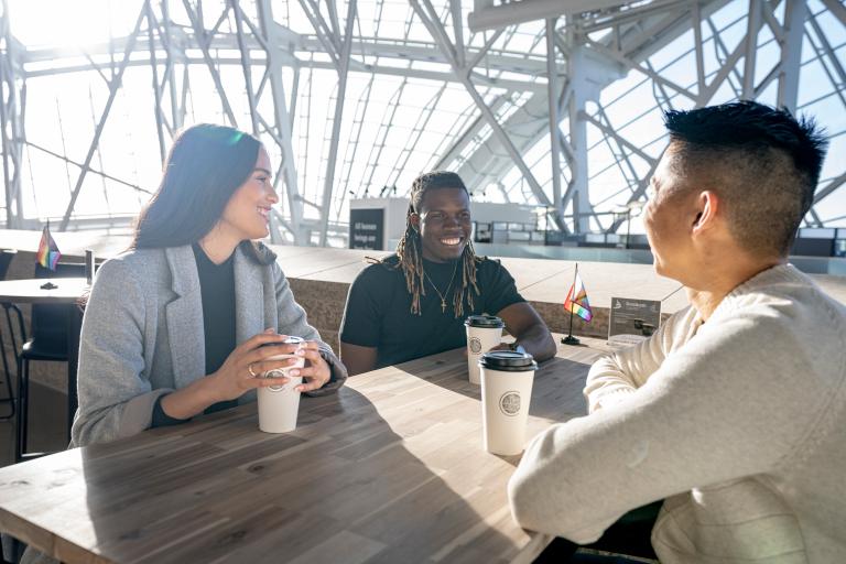 Trois personnes sont assises autour d'une table, souriantes, chacune avec une tasse à café en papier devant elle. Le soleil brille derrière elles, à travers les vitres qui entourent le Musée canadien pour les droits de la personne. Visibilité masquée.