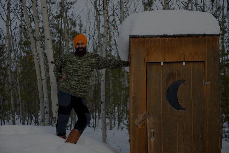 Un homme sikh-canadien, souriant, portant un pull vert, un pantalon bleu et marron, des bottes et un turban orange, adossé à une cabane avec un croissant de lune. Visibilité masquée.