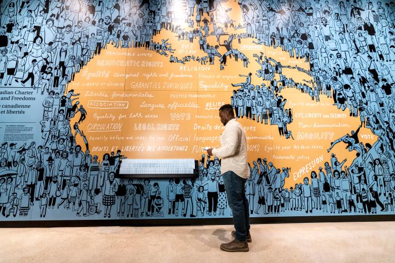 Un homme se tient devant une carte du Canada illustrée en bleu et orange. Visibilité masquée.