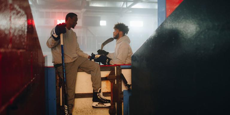 Deux personnes portant des patins de hockey et tenant des bâtons de hockey sont assises sur les bandes d’une patinoire. Visibilité masquée.