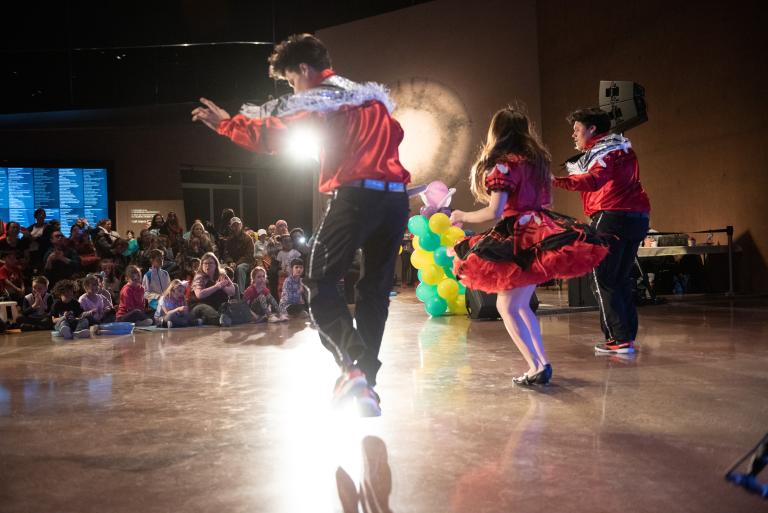 Trois danseurs habillés de rouge et de blanc se produisent devant une foule d'enfants au Musée canadien pour les droits de la personne. Visibilité masquée.