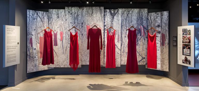 Cinq robes rouges, représentant des femmes et des filles autochtones disparues et assassinées, sont suspendues en rang dans la galerie Les parcours canadiens dans le cadre d’une exposition intitulée « Trouver de la force dans la tristesse ». Derrière les robes, la scène d’une forêt enneigée avec d’autres robes rouges accrochées aux branches des arbres est présentée sur une série de bannières. Visibilité masquée.