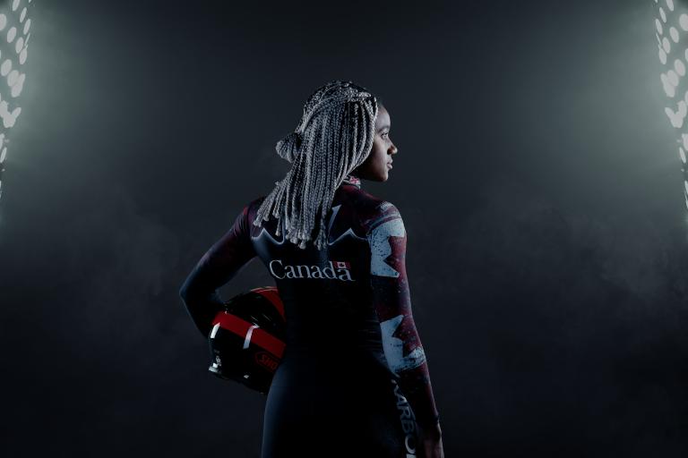 Une femme noire se tient dans un endroit sombre entre deux grands projecteurs. Elle tient un casque et porte une combinaison de bobsleigh canadienne. Visibilité masquée.