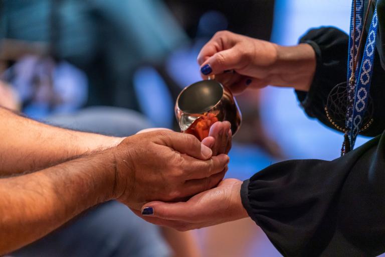 Une personne verse de l’eau d'une tasse en cuivre dans les mains d’une autre personne. Visibilité masquée.