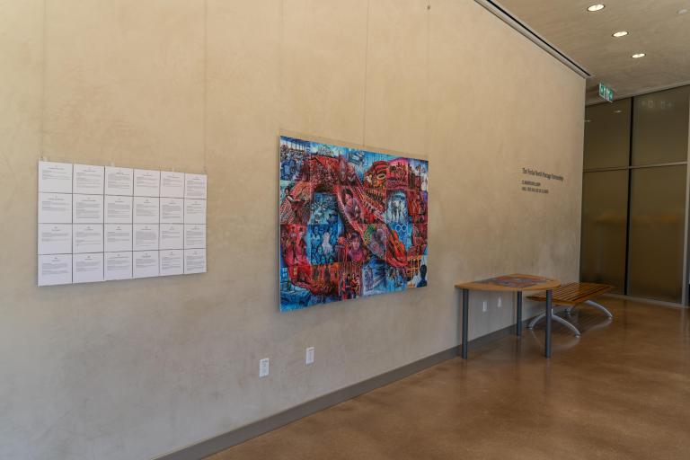 Une mosaïque de 24 tuiles sont suspendues à un mur près d’un panneau de texte. Les tuiles dépeignent des histoires sur les expériences de personnes métisses dans les systèmes scolaires coloniaux. Ensemble, les images forment un symbole de l’infini inspiré d’une ceinture fléchée rouge sur un fond bleu.