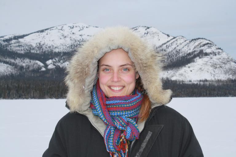 Une femme souriante portant un parka doublé de fourrure posant devant un terrain montagneux. Visibilité masquée.