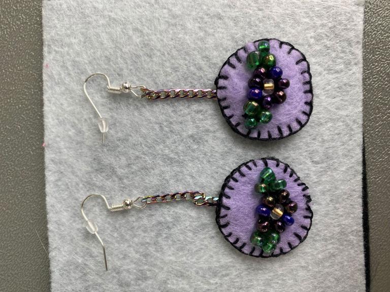 Une paire de boucles d'oreilles en feutre violet clair avec des bordures cousues violet foncé. Au milieu des boucles d'oreilles se trouvent des fleurs en perles. Visibilité masquée.