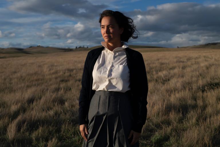 Une femme autochtone se tient dans un champ d’herbes de prairie. Derrière elle, le ciel bleu est parsemé de nuages gris et le soleil l’éclaire. Visibilité masquée.