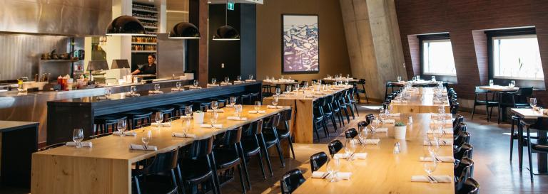 La salle à manger d'un restaurant avec de longues tables et un aperçu de la cuisine. Visibilité masquée.
