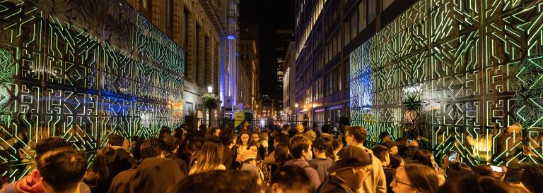 Beaucoup de gens se tiennent entre deux miroirs joints aux motifs complexes qui reflètent les lumières vertes et une foule de spectateurs. Visibilité masquée.