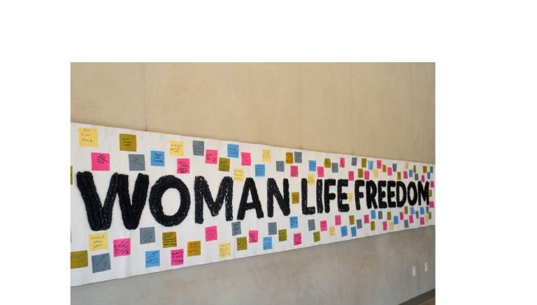 Une grande banderole blanche affiche au centre le texte anglais « Femme, vie, liberté » tissé en noir, entouré de 100 carrés de tissu colorés affichant des messages de solidarité dans différentes langues.