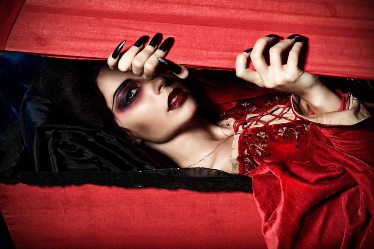 Une vampire vêtue d’une robe rouge en velours regarde à travers une boîte en bois rouge qu’elle entrouvre avec ses longs ongles noirs. Visibilité masquée.