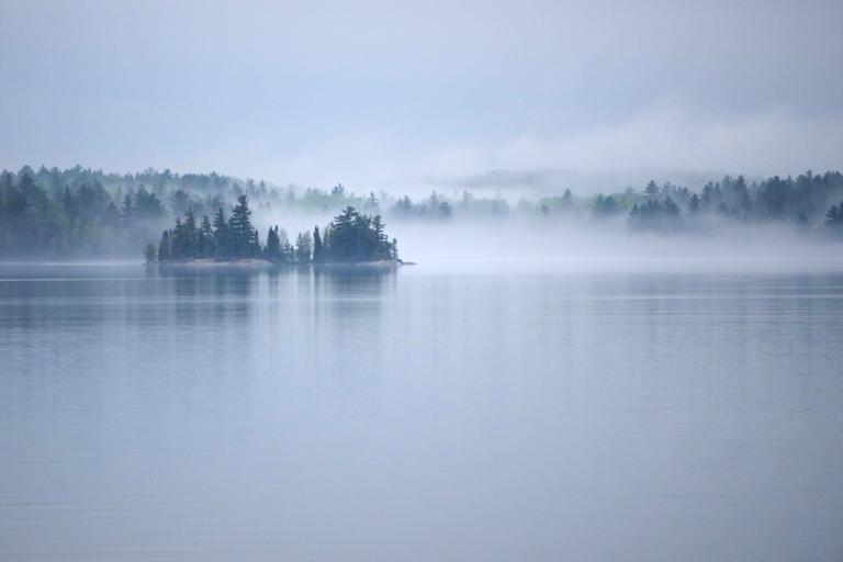 De la brume s’élève d’un grand lac calme entouré d’une forêt de conifères. Visibilité masquée.