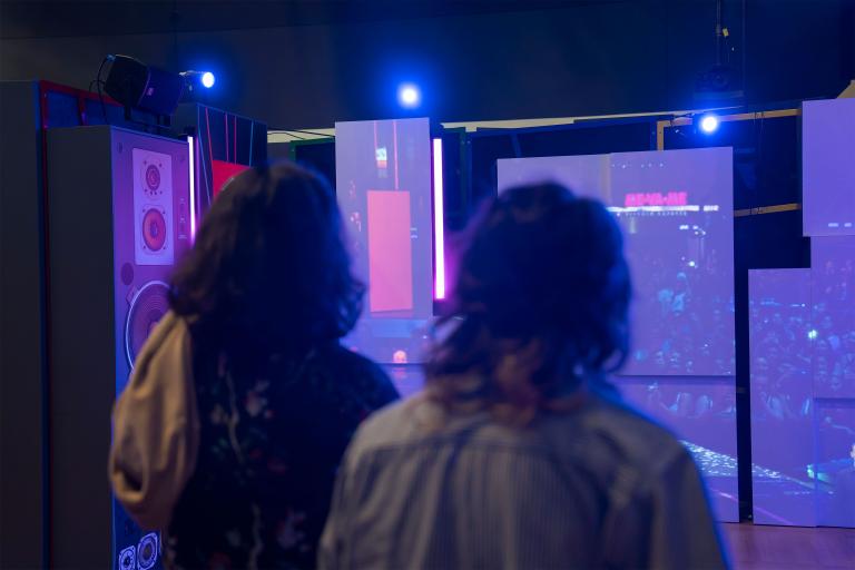 Deux personnes, vues de dos, regardent une série de panneaux d'exposition qui montrent une foule nombreuse assistant à un concert de rock. Visibilité masquée.