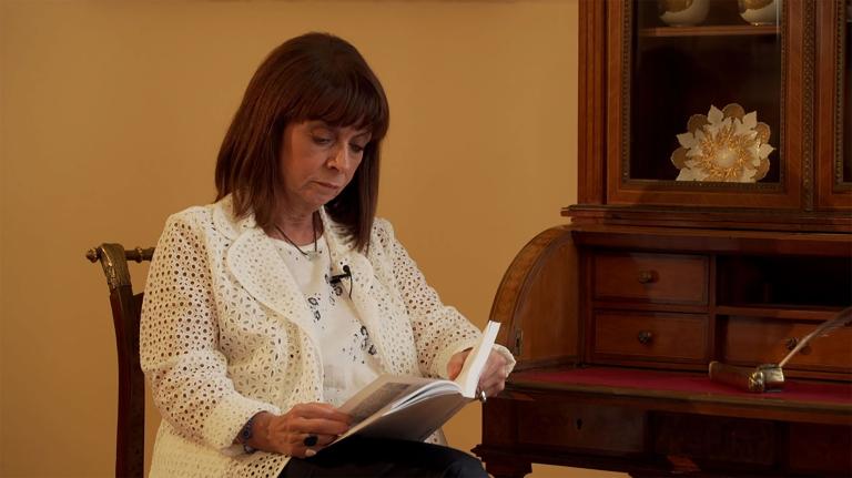 Une femme est assise en train de lire un livre. Il s’agit de Katerina Sakellaropoulou, présidente de la République hellénique. Visibilité masquée.