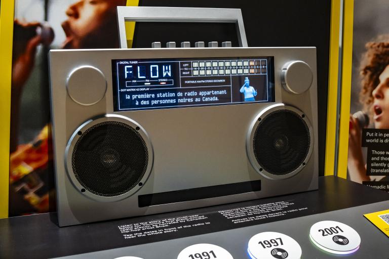 Grande radio de type "boombox" avec un écran en haut qui affiche "FLOW", des sous-titres et un interprète ASL. Visibilité masquée.