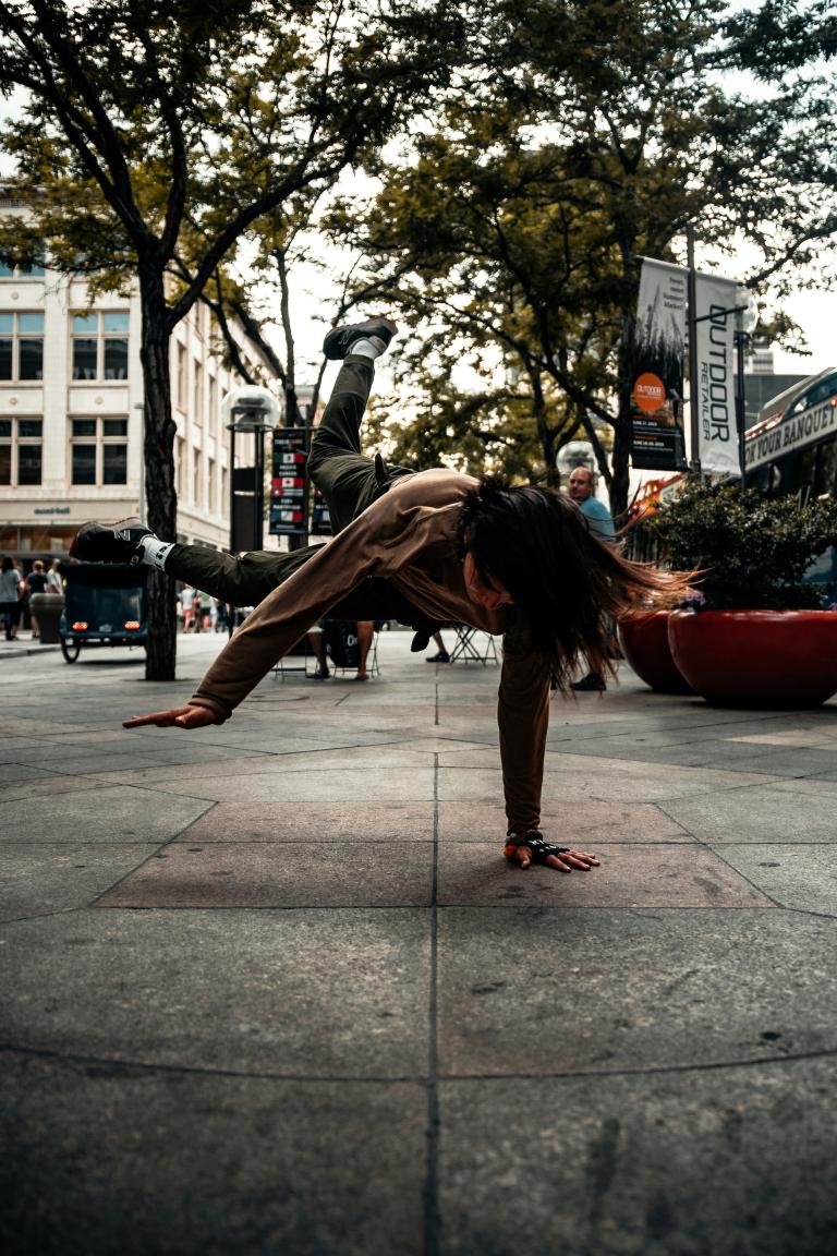 Une femme vêtue d'un coton ouaté gris et d'un pantalon gris foncé fait du breakdance sur un trottoir pendant la journée. Des arbres et des bâtiments sont à l'arrière-plan. Visibilité masquée.
