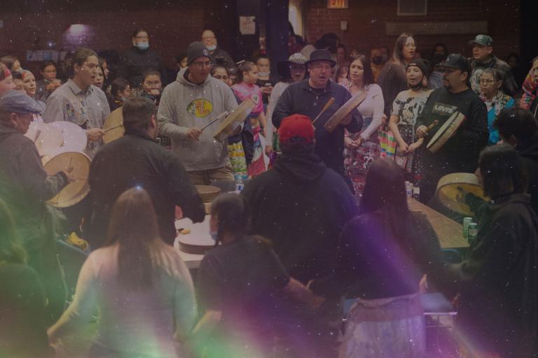 Un groupe d’hommes placés en cercle joue du tambour et chante. Une foule nombreuse et diversifiée les entoure de près et les accompagne en chantant. Visibilité masquée.