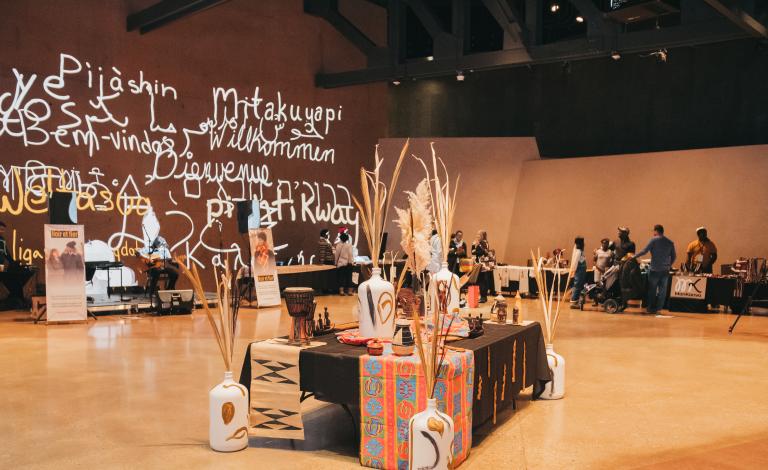 Quelques personnes se trouvent dans une salle entourée de kiosques. Au centre de la pièce, il y a une longue table ornée de nappes vibrantes et de vases remplis de longues plantes sèches. Visibilité masquée.