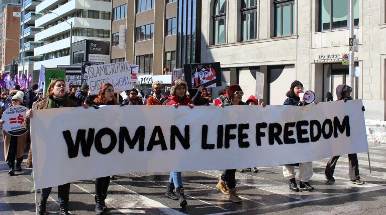 Six personnes portent une longue bannière blanche sur laquelle on peut lire les mots anglais en lettres majuscules noires "FEMME VIE LIBERTÉ" dans une rue de la ville de Toronto. Derrière elles, un défilé de femmes porte d'autres pancartes de protestation pour la Journée internationale des femmes. Visibilité masquée.
