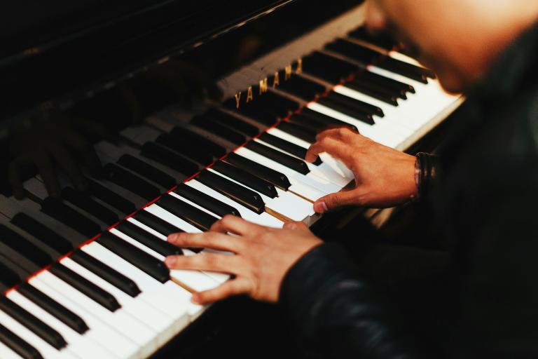 Deux mains jouent sur les touches d’un piano Yamaha d’un noir étincelant. La personne au piano, que l’on voit à peine, porte un haut noir à manches longues. Visibilité masquée.