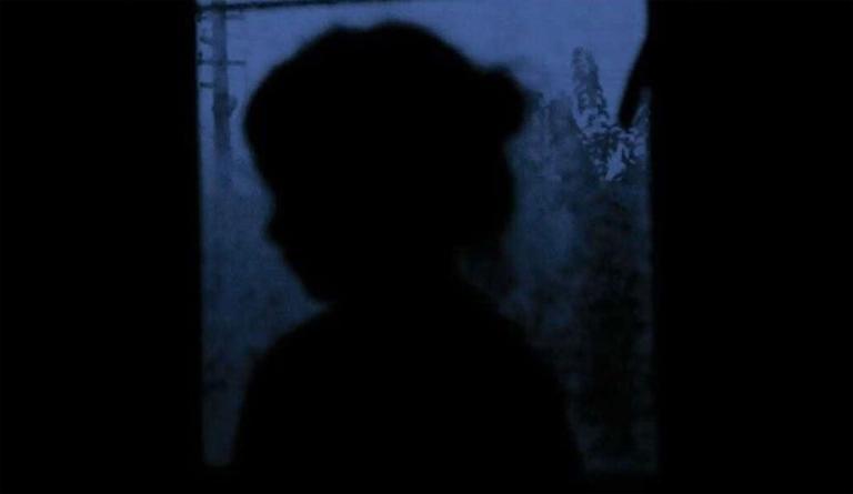 Image sombre de la silhouette d’un jeune enfant contre une fenêtre à travers laquelle on voit des arbres flous et un poteau électrique. La lumière est un ton violacé de crépuscule. Visibilité masquée.