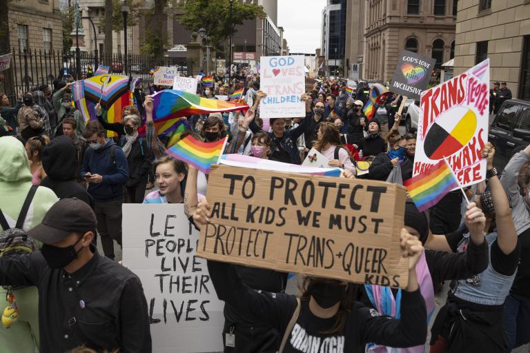Un groupe de personnes se presse dans une rue de ville en brandissant des pancartes et des drapeaux arc-en-ciel. Sur la pancarte au premier plan, il est écrit en anglais : « Pour protéger tous les enfants, nous devons protéger les enfants transgenres et queer ».
