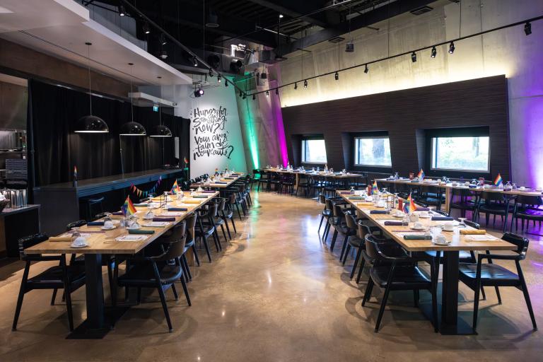 Un restaurant à aire ouverte présente des rangées de tables rectangulaires en bois recouvertes d’assiettes et de drapeaux arc-en-ciel. Visibilité masquée.