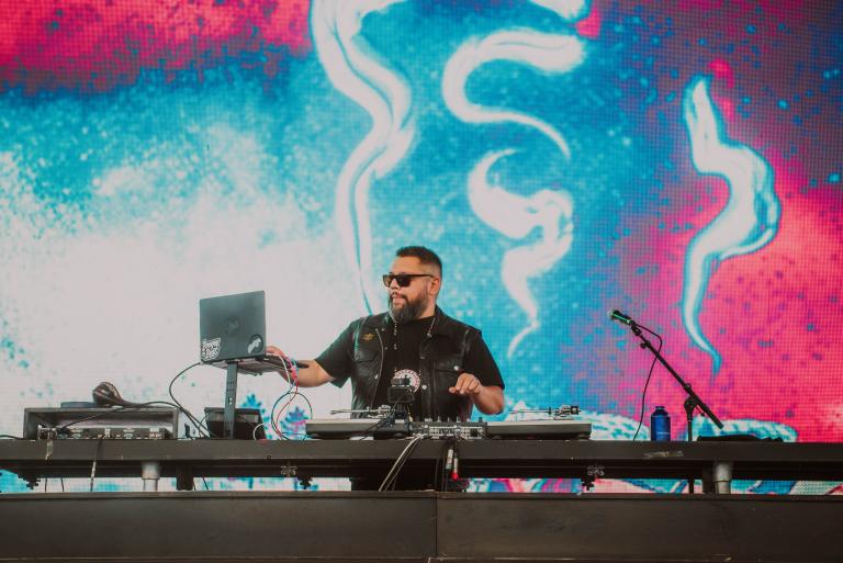 Un homme barbu aux cheveux foncés, portant des lunettes de soleil et un t-shirt noir, se produit sur scène à l’aide d’un ordinateur et d’un clavier électronique. Visibilité masquée.