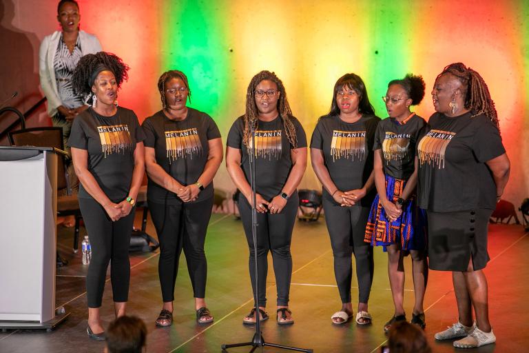 Un groupe de six artistes noires debout sur scène avec un rétroéclairage coloré. Visibilité masquée.