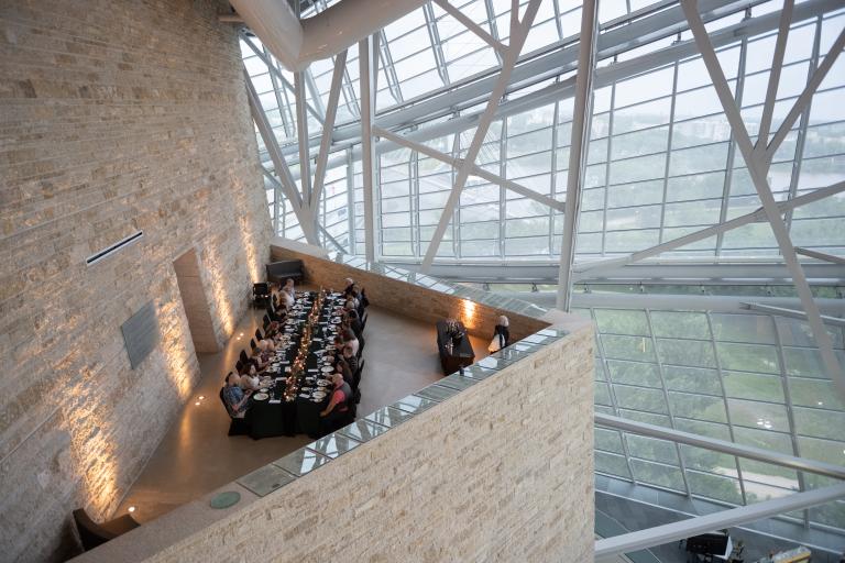 Une élégante table longue avec des personnes dînant dans un espace à l'architecture remarquable, doté de hautes fenêtres en verre et d'un plafond élevé soutenu par des poutres métalliques.