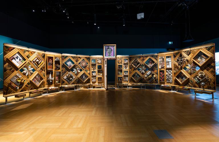 Un grand mur de bois incurvé et autoportant sur lequel sont fixés de nombreux petits objets disposés en losanges et en rectangles. Il y a une porte entrouverte au centre du mur. Visibilité masquée.