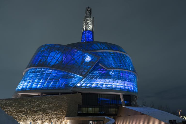 L’extérieur du Musée canadien pour les droits de la personne est photographié de nuit. Le bâtiment est éclairé de l’intérieur par des lumières vives d’un bleu profond. Visibilité masquée.