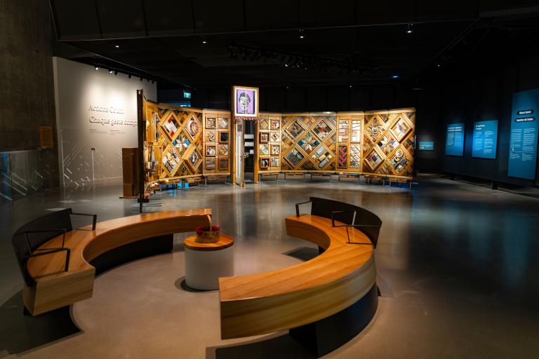 Exposition muséale présentant un grand mur de bois incurvé et autoportant sur lequel sont fixés de nombreux petits objets disposés en losanges et en rectangles. Au premier plan, des bancs circulaires créent un espace accueillant où les gens peuvent regarder et contempler l’exposition. Visibilité masquée.