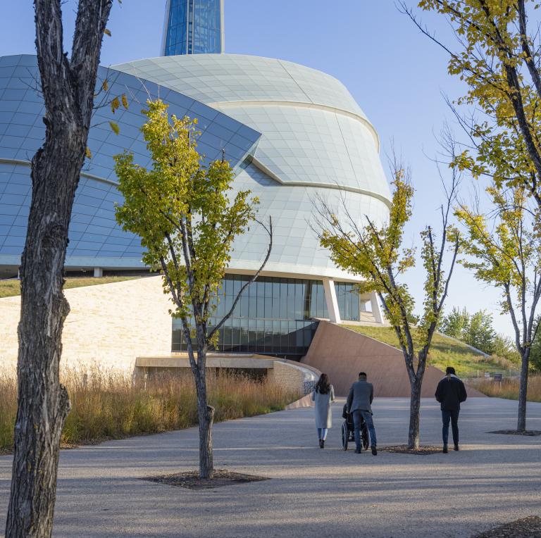 Quatre personnes suivent le chemin menant au Musée canadien pour les droits de la personne par une journée d'automne. Trois d'entre elles sont à pied et une personne est en fauteuil roulant. Visibilité masquée.
