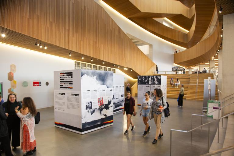 Vue intérieure d’un atrium blanc avec des niveaux en bois au-dessus. Les gens passent devant trois présentoirs d’information autoportants. Les présentoirs contiennent du texte et des images, avec pour toile de fond une architecture en bois et un escalier. Certaines personnes sont en train de discuter, tandis que d’autres interagissent avec l’exposition.
