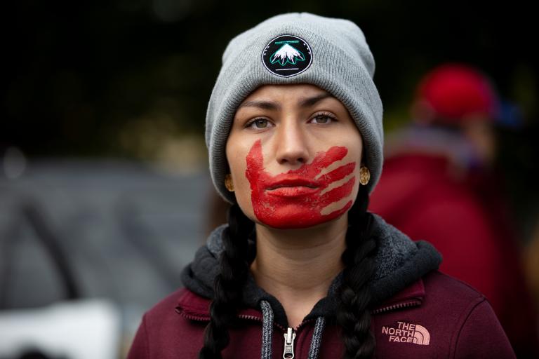 Une jeune femme aux cheveux tressés, vêtue d’une veste bordeaux et coiffée d’un bonnet d’hiver gris, regarde résolument vers l’avant, une empreinte de main rouge peinte sur la bouche et le visage. Visibilité masquée.