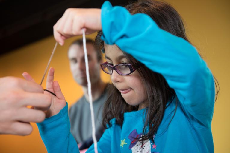 Une enfant en train de faire un bricolage travaille intensément pour tirer une ficelle. L’enfant est vêtue d'un chandail bleu orné d’un bonhomme de neige. Visibilité masquée.