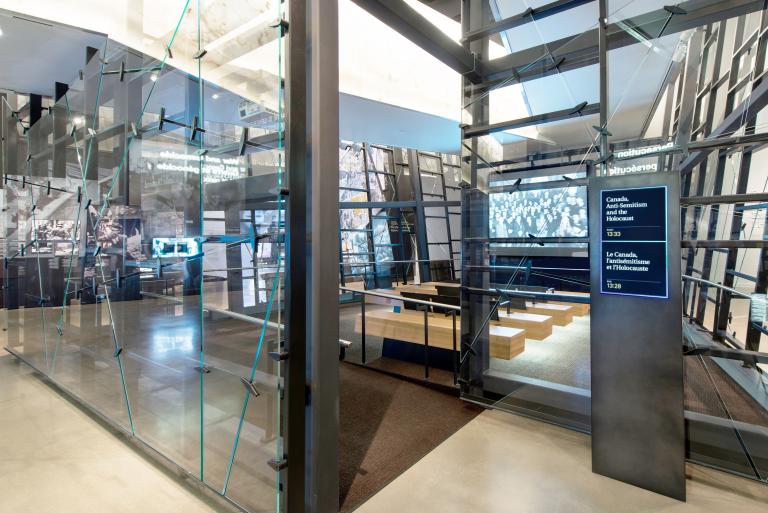 Des parois de verre inégales entourent une salle de projection avec un écran et 4 bancs en face. À l’extérieur de l’entrée de la salle, un panneau indique le film sélectionné. Visibilité masquée.