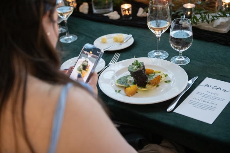 Une femme à la peau claire et aux longs cheveux bruns utilise son téléphone portable pour prendre une photo d’une assiette de nourriture gastronomique posée sur une table. Visibilité masquée.