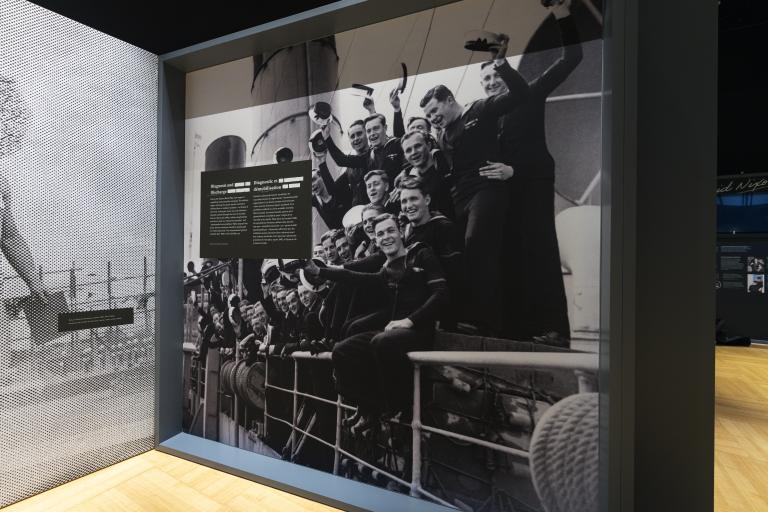 Dans une vitrine de musée, une grande photo en noir et blanc montre un groupe de marins souriant à l'appareil photo. Visibilité masquée.