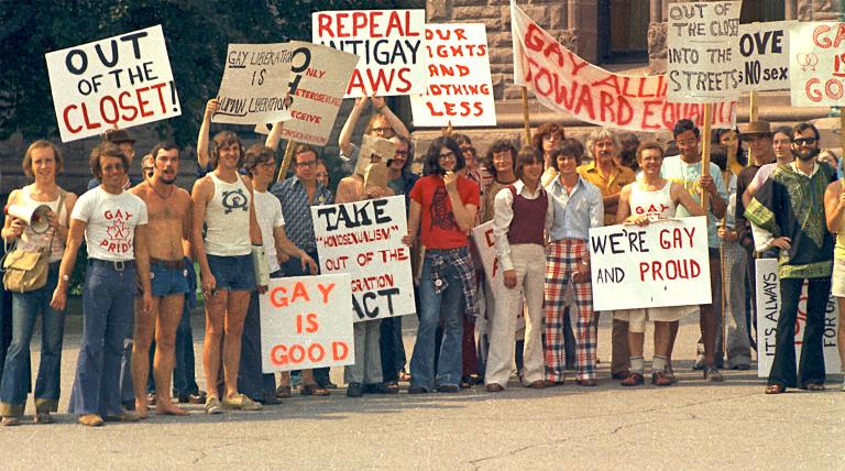 Un grand groupe de personnes vêtues de t-shirts et de pantalons évasés à la mode des années 1970 sont debout ensemble et tiennent des pancartes portant des messages en anglais tels que « sortis du placard », « c’est bien d’être gai » et « abrogez les lois anti-gaies ». Visibilité masquée.