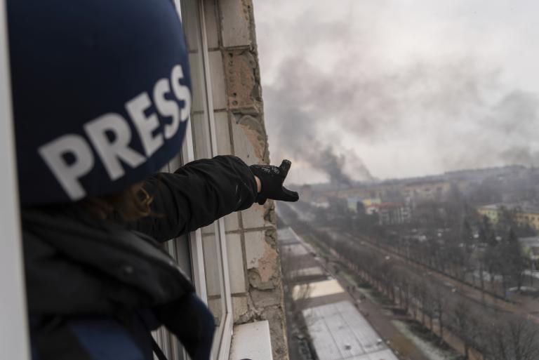 Une personne portant un casque bleu foncé marqué du mot « PRESS » montre, depuis la fenêtre d’un balcon surplombant une route, une colonne de fumée à la suite d’une explosion. Visibilité masquée.